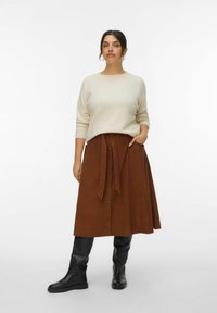 Crème gebreide trui en een bruine suede midi rok met een gestrikt middel, voorzien van knopen aan de voorkant en zijzakken. Zwarte kniehoge laarzen.