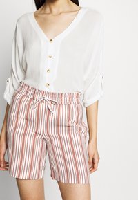 Bílá košile na knoflíky s výstřihem do V a tříčtvrtečními rukávy, spárovaná s červenobílými pruhovanými šortkami se strečovým pasem a šňůrkou.