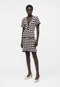 Femme debout portant une robe courte à manches courtes, longueur genou, à motifs zigzag noir et blanc avec des bords festonnés et des talons blancs ornés de nœuds.