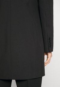 En person klädd i en svart skräddarsydd kavaj och matchande byxor, med en närbild på den nedre ryggen och högerarmen som visar fyra knapphål på ärmen.