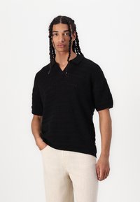 Polo shirt negru tricotat, cu decolteu în V, mâneci scurte și un model texturat. Purtați-l cu pantaloni de culori deschise, cu mâinile în buzunare.