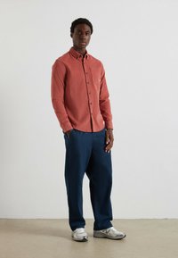 Camicia rossa a maniche lunghe con bottoni anteriori e tasca; abbinata a pantaloni blu scuro e scarpe da ginnastica grigie. Tessuto liscio, vestibilità comoda.