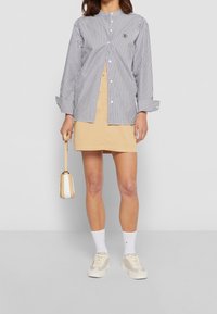 Gestreiftes Hemd mit Kragen, beigefarbener Minirock, weiße Sneaker und eine kleine beigefarbene Handtasche mit weißen Akzenten.