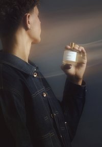 Klart glass parfymeflaske med gullkork, merket "DREAMER," holdt i en hånd; modell i en mørk denimjakke med kobberknappdetaljer.