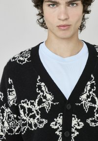 Cardigan en tricot noir avec un motif floral blanc, présentant un col en V, quatre boutons noirs à l'avant et une texture douce. Superposé sur un t-shirt bleu clair.