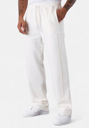 Homme portant un pantalon de survêtement blanc avec cordon de serrage à la taille, des détails de poche latérale, des baskets blanches et des bras tatoués visibles sur fond blanc.