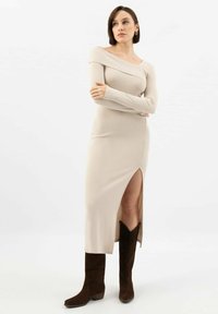 Beżowa sukienka dzianinowa z prążkowanej tkaniny, z dekoltem odsłaniającym ramiona i rozcięciem do uda, zestawiona z brązowymi za kostkę botkami. Gładka tekstura, dopasowany krój.