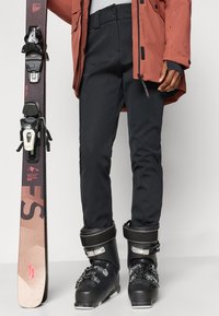 Μαύροι σκι παντελόνια με ευθεία γραμμή, συνδυασμένα με μαύρα παπούτσια σκι που διαθέτουν μεταλλικές αγκράφες. Δίπλα κρατείται ένα σκι με καφέ σχέδιο.