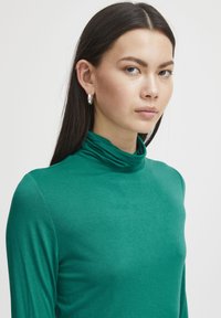 Grünes Langarm-Turtleneck-Oberteil aus einem weichen, glatten Stoff. Verfügt über ein figurbetontes Design mit einer dezenten Nackenraffung. Silberne, kreisförmige Ohrringe.