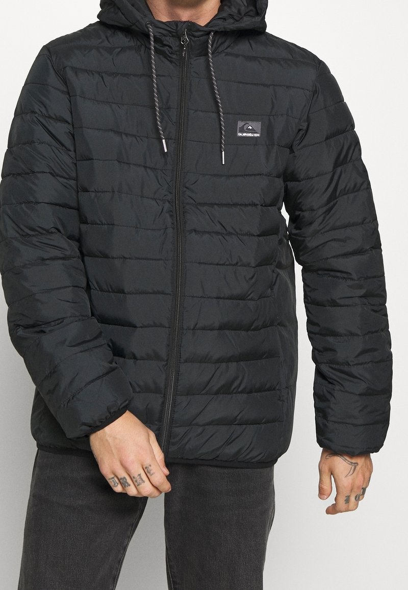 Veste noire matelassée avec un design en quilting, dotée d'une capuche, d'une fermeture éclair frontale et d'un cordon de serrage, fabriquée en tissu lisse et léger.