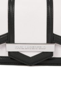 Fehér bőr kézitáska fekete akcentusokkal, geometriai mintázati elemekkel és fémből készült táblával, amelyen a "KARL LAGERFELD" felirat található.