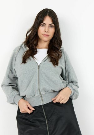 Jeune femme aux longs cheveux foncés portant un sweat à capuche gris zippé par-dessus un haut blanc et une jupe noire avec une fermeture éclair à l'avant.