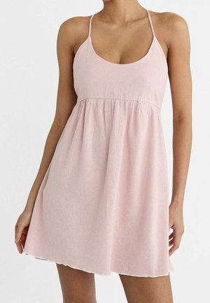 Kobieta ubrana w bezrękawnikową, jasno-różową, prążkowaną mini sukienkę dzianinową z cienkimi ramiączkami i marszczonym stanem.