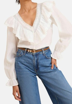 Femme portant un chemisier blanc à manches longues avec un col volanté en dentelle et un jean bleu taille haute avec une ceinture beige, main posée sur la hanche.