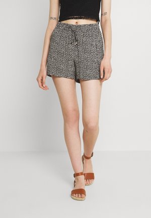 Mujer vistiendo un top corto negro y pantalones cortos florales en blanco y negro con sandalias marrones de tiras en el tobillo, de pie frente a un fondo blanco.