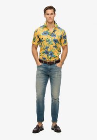 Camisa amarilla de manga corta con patrón floral azul, combinada con jeans azul claros y mocasines marrones. Presenta un diseño de botones.