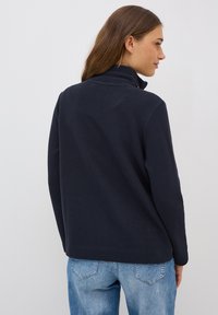 Marineblauer Pullover aus strukturiertem Stoff mit hohem Kragen und langen Ärmeln, kombiniert mit hellblauen Jeans. Rückansicht dargestellt.