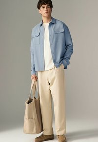 Camisa de botones azul claro con dos bolsillos en el pecho, camiseta crema por debajo, pantalones beige y una bolsa de mano marrón claro, combinada con zapatos marrones.