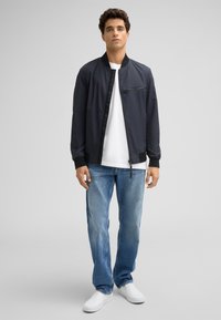Marineblaue Bomberjacke mit glatter Textur, ausgestattet mit einer Reißverschluß-Brusttasche. Kombiniert mit hellblauen geraden Jeans und weißen Schuhen.