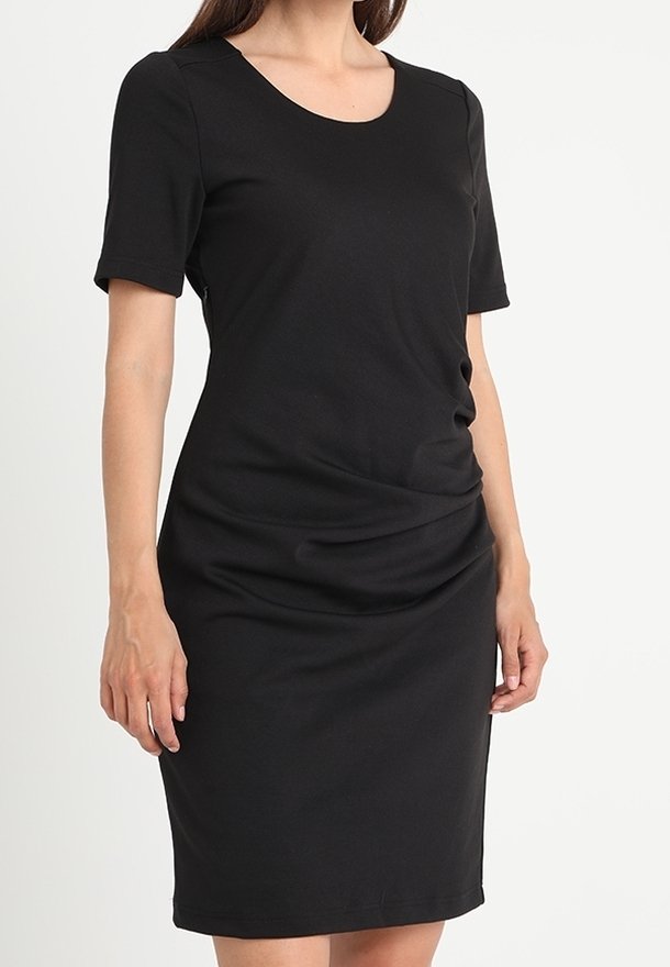 Femme portant une robe noire moulante à manches courtes et longueur genou avec un léger froncé à la taille sur un fond uni.