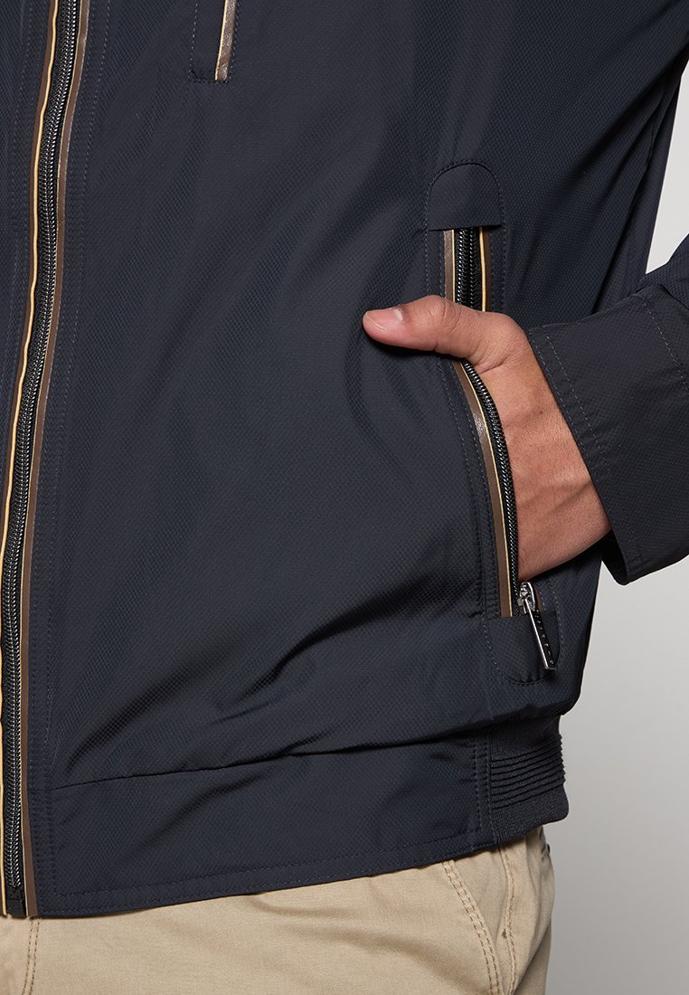 Marineblau leichte Jacke mit strukturiertem Obermaterial, goldenen Reißverschlüssen und Seitentaschen; imta sich durch einen enganliegenden Schnitt mit elastischen Bündchen aus.