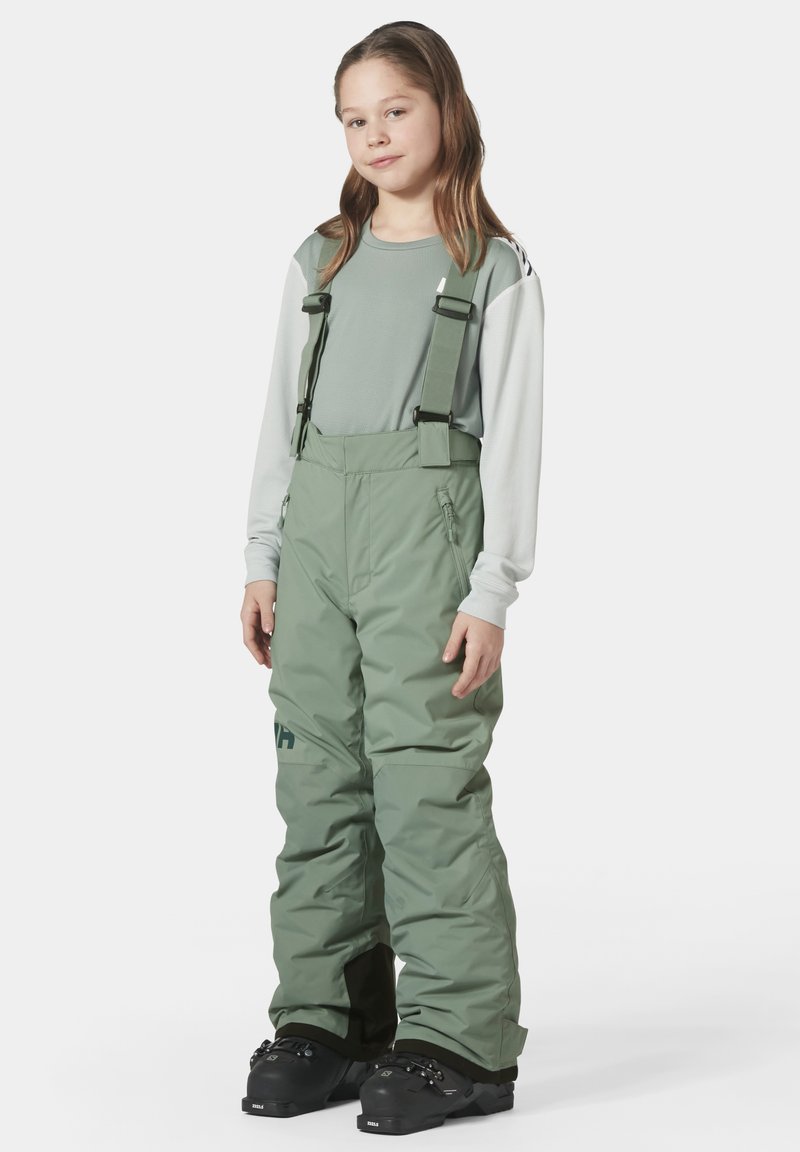 Pantalones de nieve verdes aislados con tirantes ajustables, bolsillos con cremallera y un corte holgado. Combinados con una camiseta de manga larga gris claro.