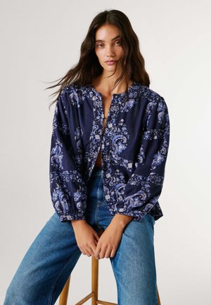 Kvinde med langt mørkt hår iført en marineblå blomsterbluse med pufærmer og blå jeans, siddende på en træskammel mod en hvid baggrund.