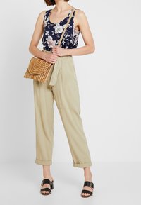 Blumiges navyblaues Tanktop kombiniert mit hellbeigen, hochgeschnittenen Hosen, einer geflochtenen, braunen Umhängetasche und Sandalen mit schwarzen Riemen.