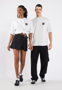 Two individuals wear matching white t-shirts featuring black graphic prints. One pairs a knotted tee with a black skirt; the other has black pants.