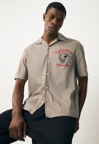 Chemise boutonnée à manches courtes, tissu gris clair, motif brodé featuring un oiseau et le texte "Yellowstone National Park" en rouge.