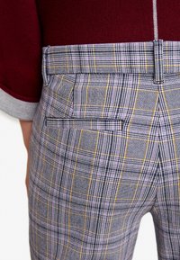 Gros plan sur l'arrière d'un pantalon à carreaux gris avec des lignes jaunes et violettes, poche passepoilée, passant de ceinture, porté avec un haut en maroon et gris à revers.