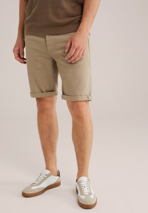 Uomo che indossa pantaloni corti beige arrotolati e sneakers bianche, in piedi su uno sfondo chiaro e uniforme.