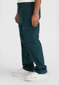 Teal corduroy cargobroek met zijzakken, een relaxte pasvorm en een zachte textuur. Gecombineerd met witte sneakers voor contrast.