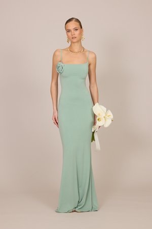 Femeie purtând o rochie mulată verde pal cu bretele și detaliu floral pe piept, ținând crini albi, stând în fața unui fundal simplu bej.