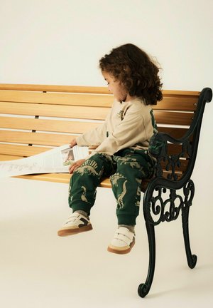 Bambino con capelli ricci seduto su una panchina di legno che legge un giornale, indossando pantaloni verdi con stampa di dinosauri e sneakers beige.