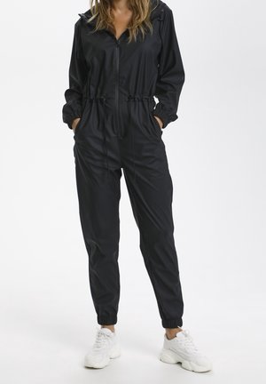Kvinde iført en sort zip-up jumpsuit med elastiske manchetter og hvide chunky sneakers, stående med hænderne i lommerne på en hvid baggrund.