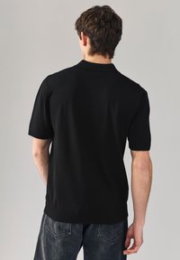 Polo noir en maille avec manches courtes, col et ourlet côtelés. Présente une texture unie, une coupe ajustée et un design minimaliste.