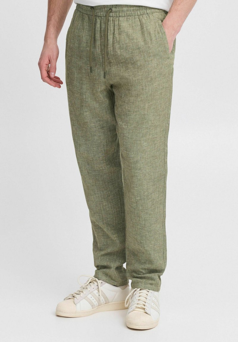 Homme portant un pantalon en lin vert clair avec taille élastique et cordon, associé à des baskets blanches et beiges, debout devant un fond uni.