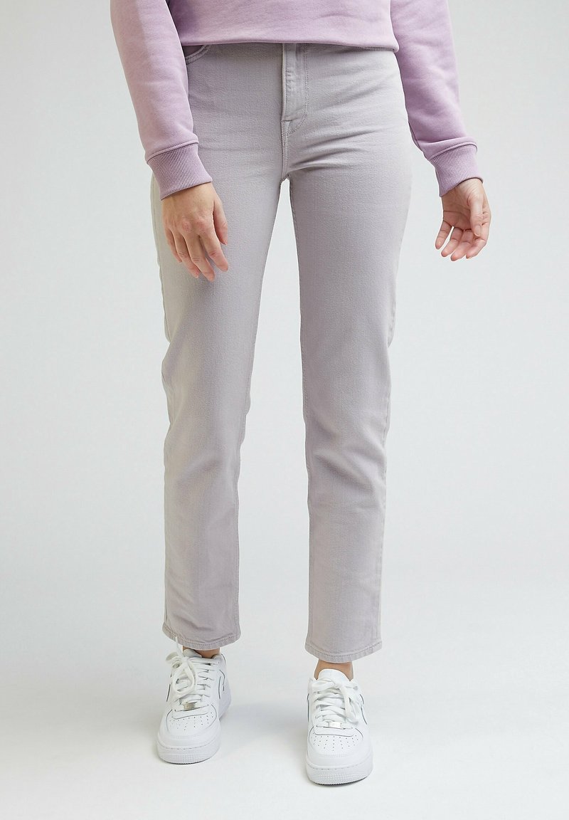 Personne portant un pantalon gris clair droit, des baskets blanches et un haut à manches longues violet clair, debout devant un fond uni.
