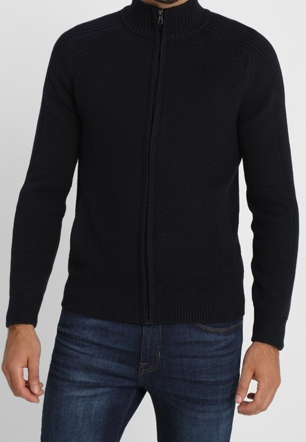 Tmavě modrý svetr na zip s vysokým límcem, který má strukturovaný pletený vzor a ribbované manžety a spodní lem. Nosený přes modré džíny.