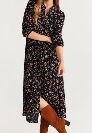 Kobieta ubrana w czarną, kwiatową sukienkę midi z rękawami do łokcia oraz brązowe zamszowe kozaki za kolano, stojąca z założonymi skrzyżowanymi ramionami.