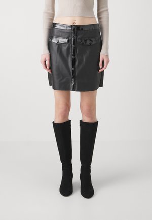 Gonna mini in pelle nera con dettagli lucidi, chiusura frontale a bottoni, due tasche a patta e dettagli con spacchi laterali. Abbinata a stivali neri al ginocchio.