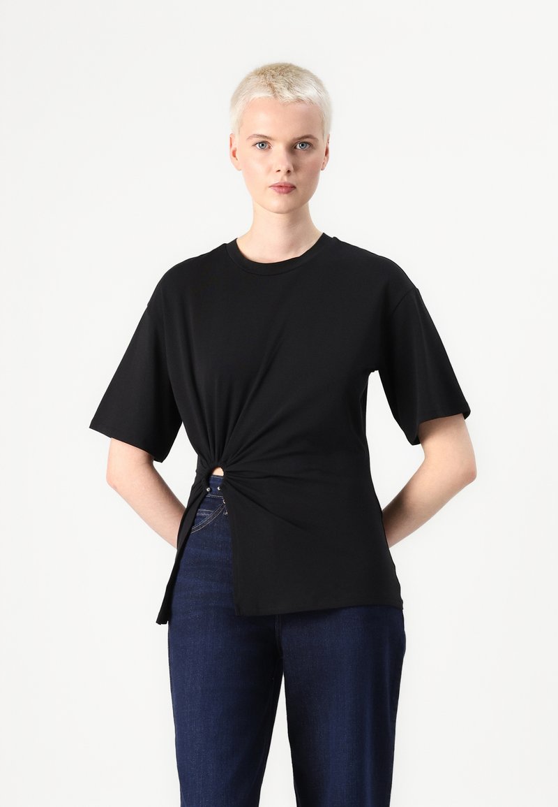 Czarny t-shirt z krótkim rękawem z detalem w postaci węzła z przodu, o luźnym kroju i miękkim materiale, zestawiony z ciemnymi dżinsami.