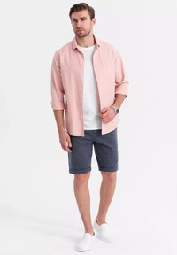 Chemise boutonnée rose clair, T-shirt blanc, shorts navy avec ourlet retourné, baskets blanches, debout sur un fond uni.