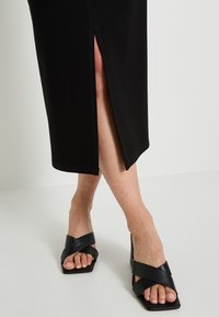 Robe noire, à mi-mollet, avec une fente sur le côté. Portée avec des sandales en cuir noires dotées de lanières croisées et de bouts carrés.