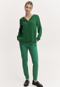 Green blouse with a V-neck and buttons, paired with matching green slim-fit pants. Both made of smooth fabric. Model wears black loafers.