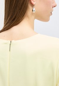 Γυναίκα με ίσια ανοιχτό καστανά μαλλιά, που φοράει ανοιχτό κίτρινο τοπ με φερμουάρ στο πίσω μέρος και ασημένιο σκουλαρίκι σε σχήμα δάκρυ στο αριστερό αυτί.