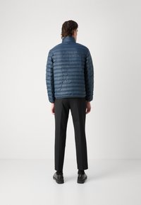 Giacca puffa blu navy con design a rombi orizzontali, colletto alto e finitura liscia; indossata con pantaloni e scarpe neri.