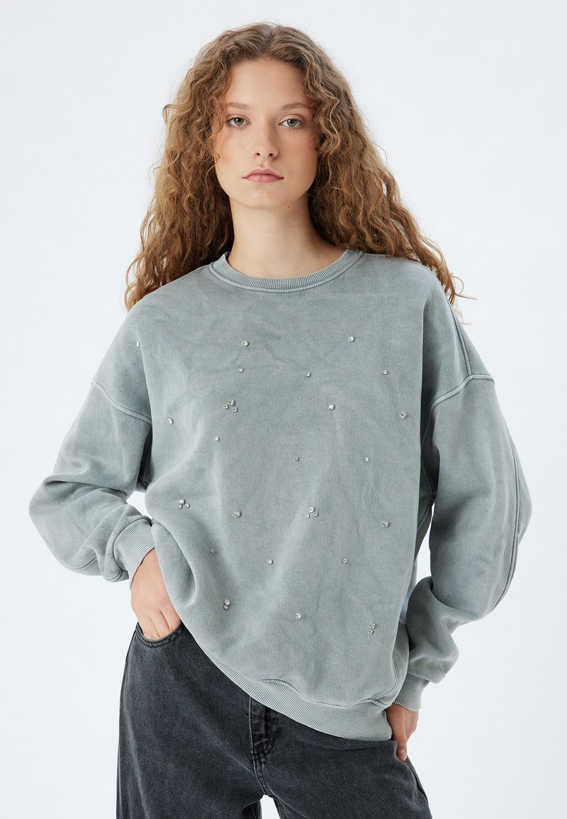 Ljusgrå sweatshirt gjord av mjukt material, prydd med spridda rhinstenar på framsidan. Den har rund hals och avslappnade ärmar, i kombination med mörka jeans.