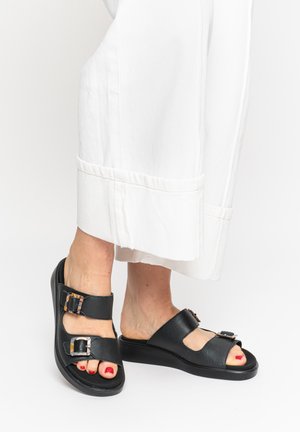 Füße in schwarzen offenen Sandalen mit doppelten Schnallen, kombiniert mit weißen Hosen mit weitem Bein vor einfachem Hintergrund.